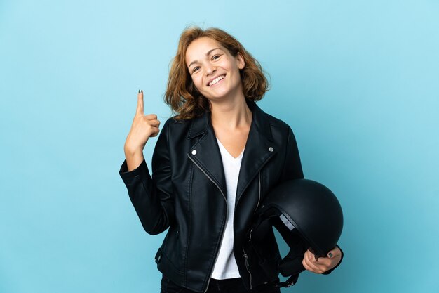 Chica georgiana sosteniendo un casco de motocicleta aislado en la pared azul mostrando y levantando un dedo en señal de lo mejor