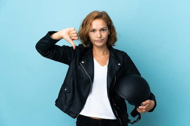 Chica georgiana sosteniendo un casco de motocicleta aislado sobre fondo azul mostrando el pulgar hacia abajo con expresión negativa