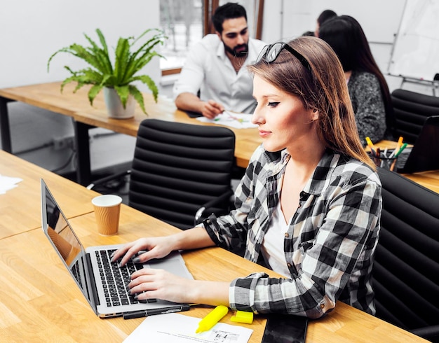 Foto chica gerente en camisa casual a cuadros que trabaja en la computadora portátil en interiores en una acogedora oficina blanca