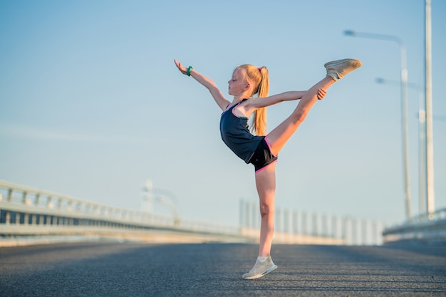 Chica gimnasta comprometida en el verano en la calle, sobre un fondo de cielo azul, cordeles, estiramiento, arabescos