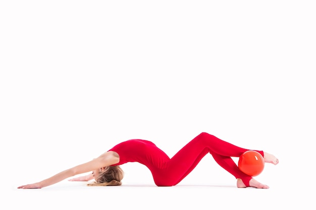 Chica gimnasta en un mono rojo hace un ejercicio con una pelota sobre un fondo blanco.