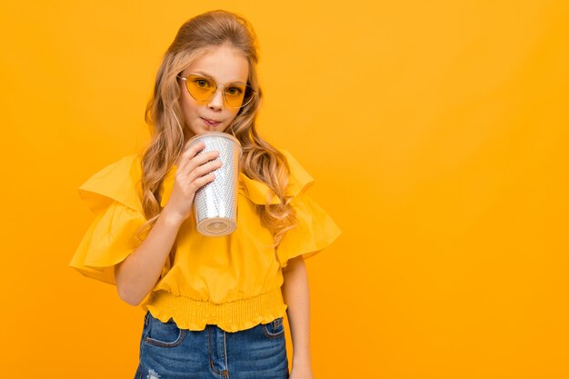 Chica glamorosa en gafas brillantes bebe un cóctel en una pared amarilla