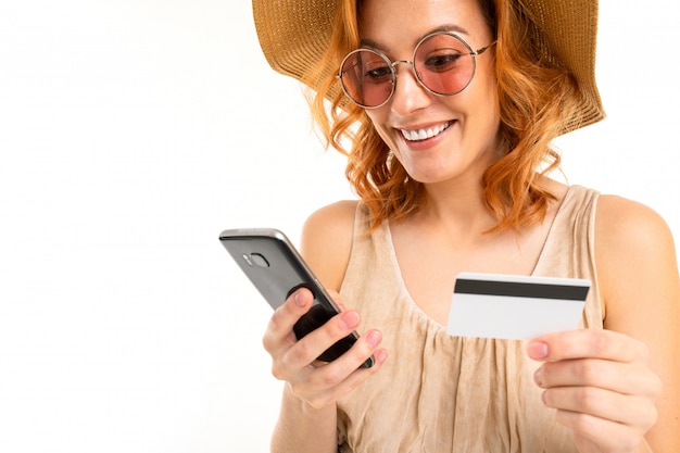 Chica glamorosa con un sombrero vestido con un vestido de verano y gafas de sol pide boletos por teléfono y tiene una tarjeta de crédito en una pared blanca