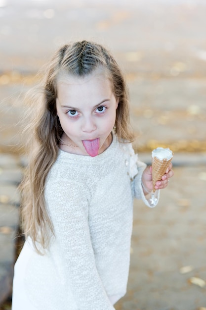 Chica golosa en alegre mueca con lengua come helado, fondo claro. Niña niño con cono de helado en la mano. Concepto de golosinas de verano. Niño goloso con helado blanco en cono de galleta.