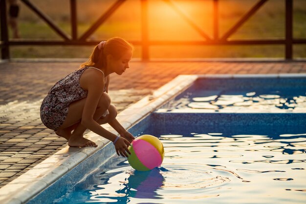 Una chica gorda y bronceada con el pelo oscuro trenzado en un moño con un brillante traje de verano está jugando cerca de la piscina con una pelota inflable en el fondo del sol dorado de la tarde de verano