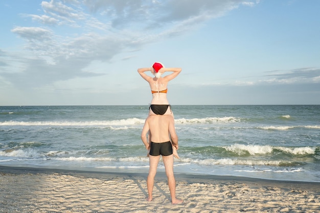 Chica con gorro de Papá Noel se sienta sobre los hombros de un chico Vista trasera de la orilla del mar