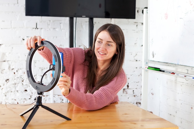 Foto chica grabando video en un teléfono inteligente y encendiéndose con una lámpara de anillo en la mesa en la sala de luz