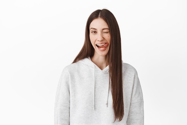 Una chica graciosa y positiva te muestra la lengua y te guiña el ojo sonriendo ampliamente de pie, juguetona y feliz disfrutando de momentos despreocupados posando con una sudadera con capucha gris informal contra un fondo blanco