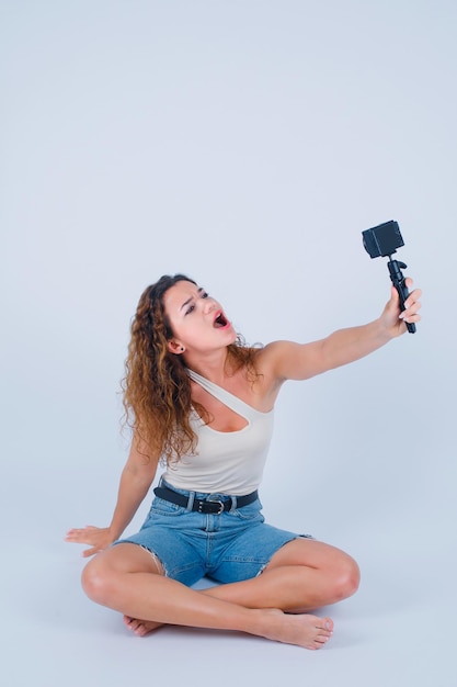 Chica gritando está tomando selfie con su mini cámara sentada en el suelo sobre fondo blanco.