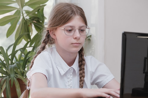 Chica guapa adolescente escribiendo en una computadora portátil y haciendo los deberes sentado detrás de la mesa en casa. Hermosa joven con gafas, aprendizaje en línea con una computadora portátil, educación a distancia, autoeducación