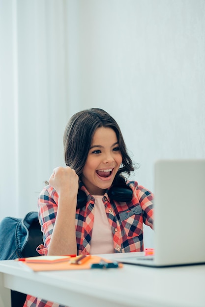 Chica guapa alegre sentada en la mesa con una computadora portátil frente a ella y sonriendo mientras mira la pantalla y celebra la victoria