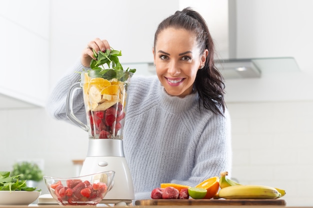 Foto una chica guapa apila una colorida mezcla de frutas y hojas verdes en una licuadora en la cocina de casa.