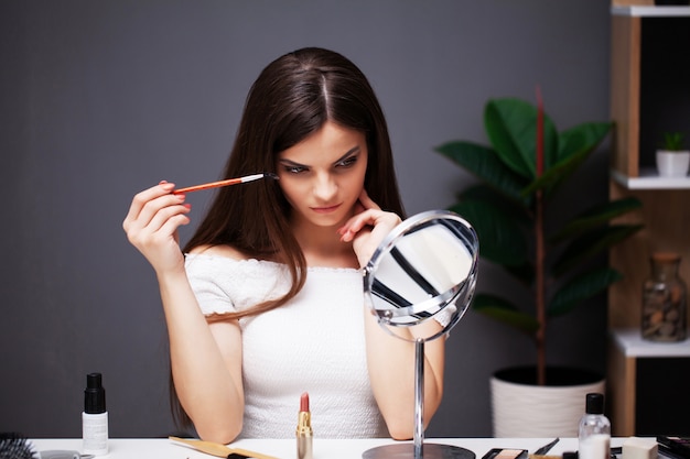 Chica guapa aplica maquillaje en la cara en casa cerca del espejo