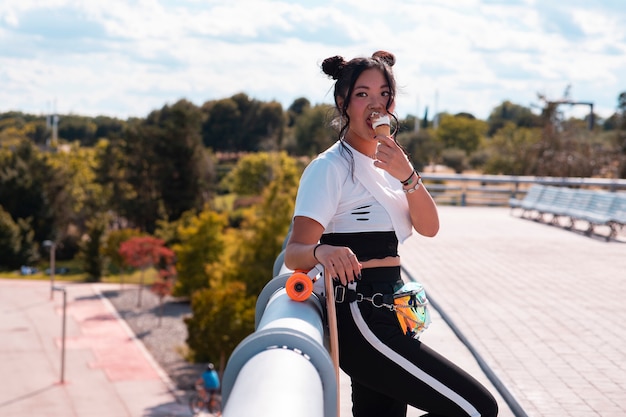 Chica guapa asiática comiendo helado en el parque, ropa casual y patineta