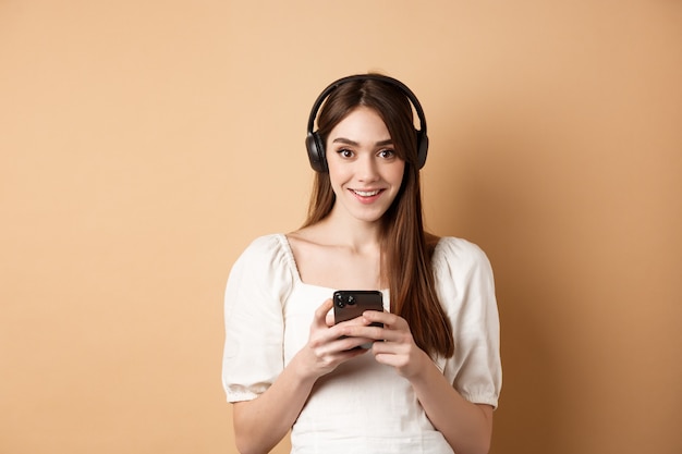Chica guapa en auriculares sonriendo a la cámara, escuchando música y usando la aplicación de teléfono móvil, fondo beige.