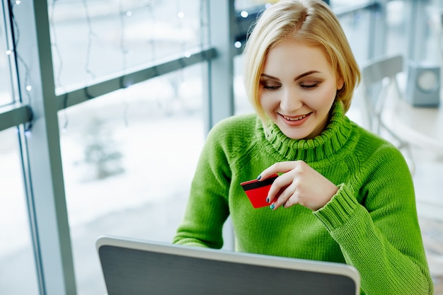 Chica guapa con cabello claro con suéter verde sentado en la cafetería con laptop, retrato, concepto independiente, compras en línea.