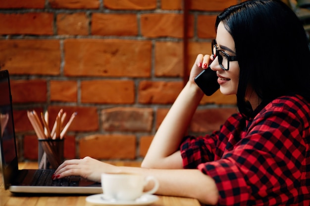 Chica guapa con cabello negro con anteojos sentado en la cafetería con computadora portátil, teléfono móvil y taza de café, concepto independiente, retrato, espacio de copia, con camisa roja.