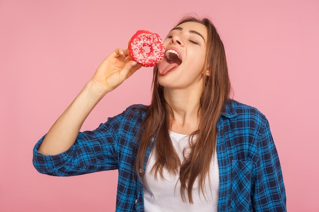 Chica guapa con camisa a cuadros de pie con los ojos cerrados y lamiendo donut dulce soñando con morder donut sabroso luchando contra la tentación de comer dulces azucarados tiro de estudio interior aislado