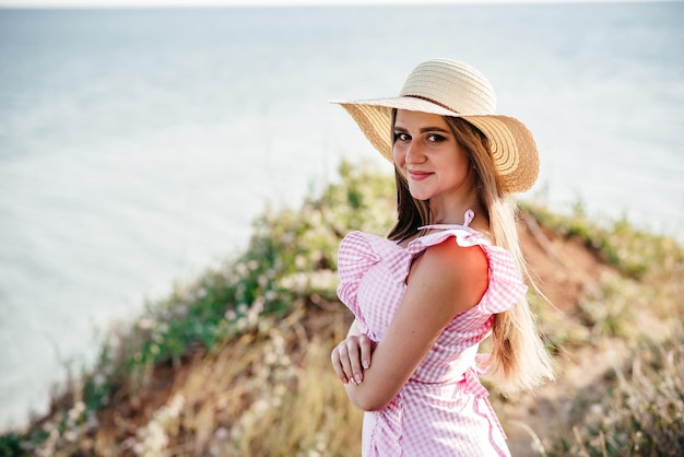 Chica guapa en el campo al atardecer Tono vintage Chica elegante caminando en un prado soleado al atardecer atmosférico Chica sosteniendo un sombrero de paja Una chica con un vestido blanco se encuentra contra el fondo de las montañas