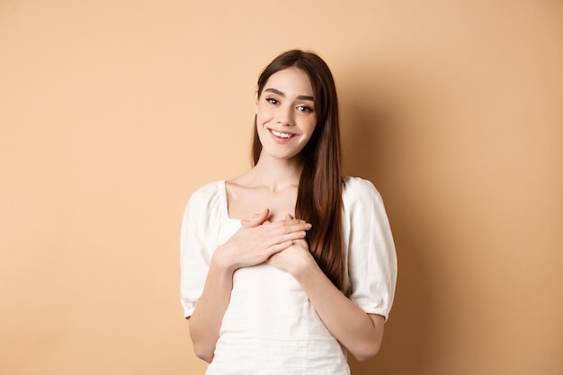 Chica guapa cogidos de la mano en el corazón y sonriendo, agradeciéndole, sintiéndose agradecida y complacida, de pie en vestido sobre fondo beige.