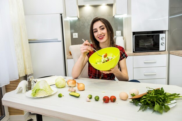 Chica guapa comiendo ensalada de verduras frescas mientras sonríe y está sentado en la cocina de su casa.