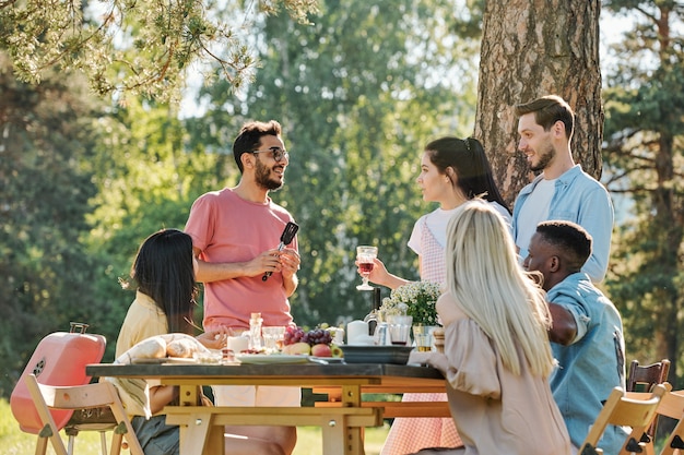 Chica guapa con una copa de vino tinto de pie junto a su novio y hablando con un amigo de raza mixta por la mesa festiva servida durante la cena al aire libre