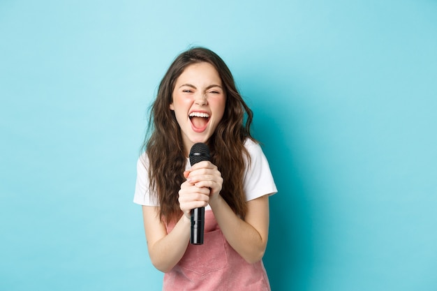 Chica guapa emocionada cantando karaoke, sosteniendo el micrófono y sonriendo feliz, de pie sobre fondo azul.