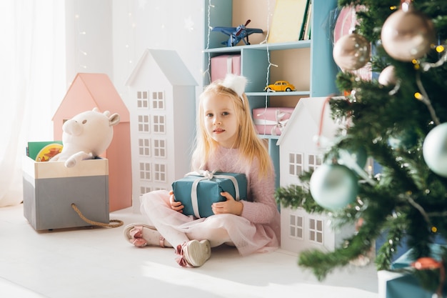 Chica guapa está sentada en el árbol de Navidad y sosteniendo una caja con un regalo