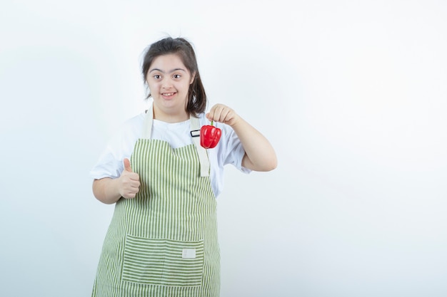 Chica guapa joven en delantal a cuadros sosteniendo pimiento rojo y mostrando el pulgar hacia arriba.