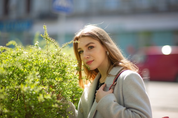 Chica guapa joven elegante con un abrigo gris mira a la cámara y sonríe levemente en el contexto de la ciudad. La niña está junto a un arbusto verde.