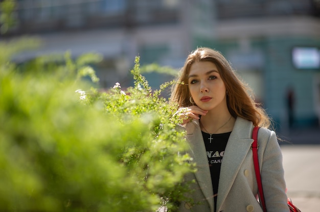 Chica guapa joven elegante con un abrigo gris mira a la cámara y sonríe levemente en el contexto de la ciudad. La niña está junto a un arbusto verde.