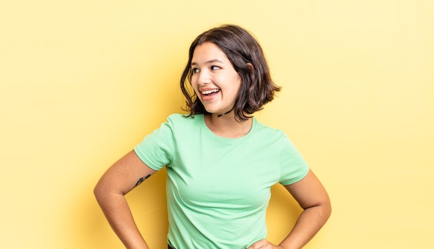 Chica guapa joven que parece feliz, alegre y segura, sonriendo con orgullo y mirando al lado con ambas manos en las caderas