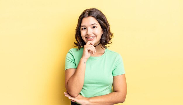 Chica guapa joven que parece feliz y sonriente con la mano en la barbilla, preguntándose o haciendo una pregunta, comparando opciones