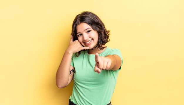 Chica guapa joven sonriendo alegremente y apuntando a la cámara mientras hace una llamada, gesto más tarde, hablando por teléfono