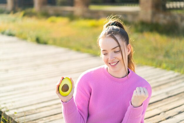 Chica guapa joven sosteniendo un aguacate al aire libre celebrando una victoria