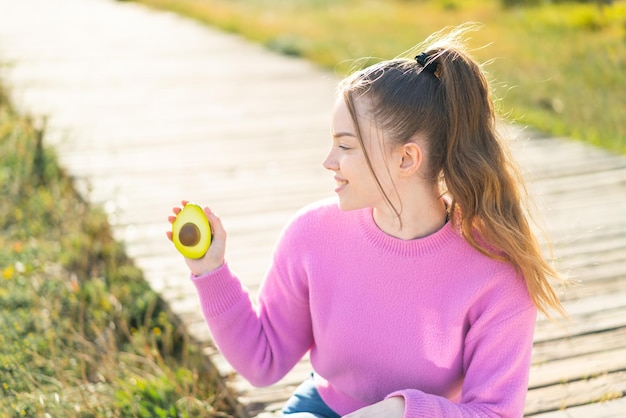 Chica guapa joven sosteniendo un aguacate al aire libre con expresión feliz