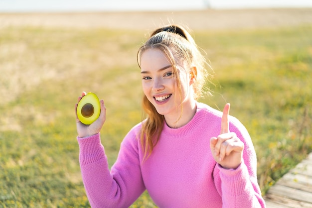 Chica guapa joven sosteniendo un aguacate al aire libre señalando una gran idea