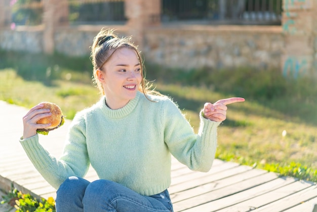 Chica guapa joven sosteniendo una hamburguesa al aire libre apuntando hacia un lado para presentar un producto