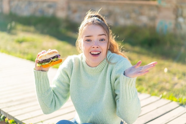 Chica guapa joven sosteniendo una hamburguesa al aire libre con expresión facial sorprendida