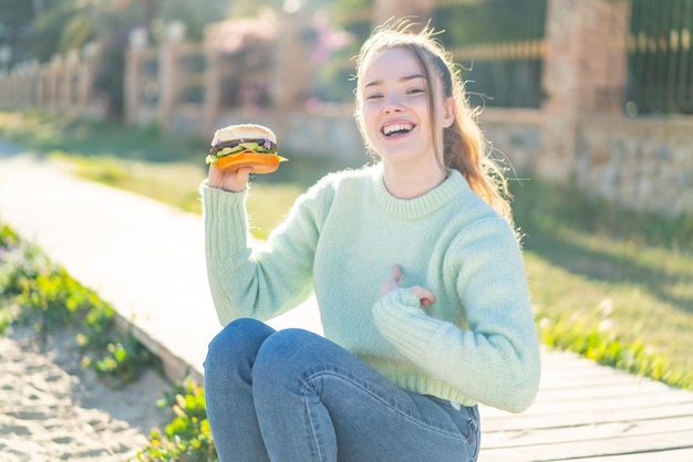 Chica guapa joven sosteniendo una hamburguesa al aire libre con expresión facial sorpresa