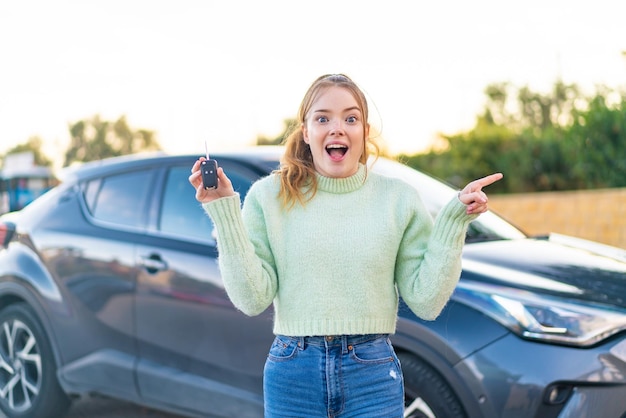 Chica guapa joven sosteniendo las llaves del auto al aire libre sorprendida y señalando con el dedo hacia un lado