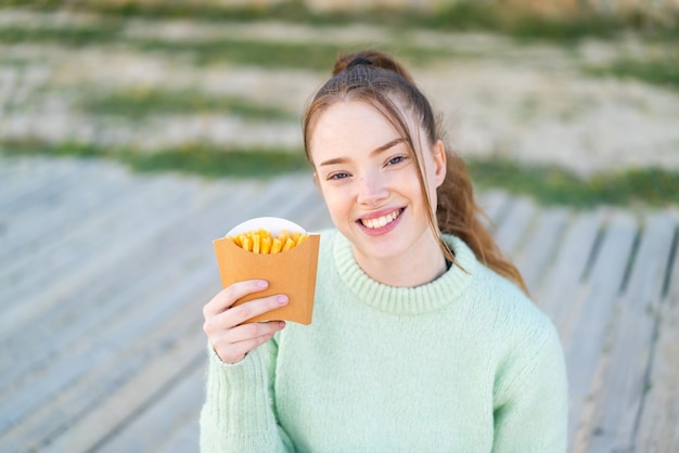 Chica guapa joven sosteniendo papas fritas al aire libre sonriendo mucho