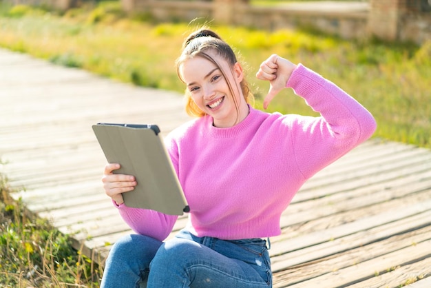 Chica guapa joven sosteniendo una tableta al aire libre orgullosa y satisfecha