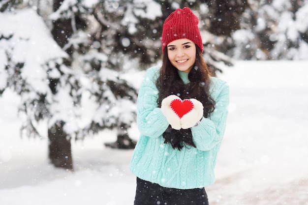 Chica guapa joven con un suéter azul cálido y guantes en el fondo de árboles cubiertos de nieve sosteniendo un corazón rojo y sonriendo. Día de San Valentín.