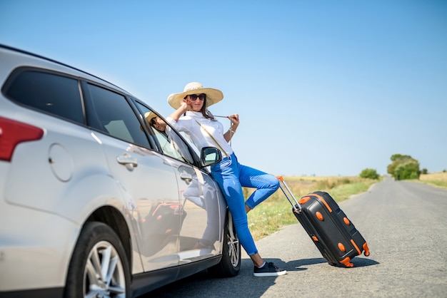 Chica guapa con maleta de pie cerca del coche y wiat para su viaje de ensueño.