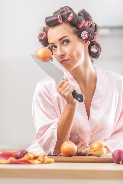 Chica guapa mira a la cámara con una cebolla clavada en el cuchillo en la mano mientras está de pie en camisón en la cocina.