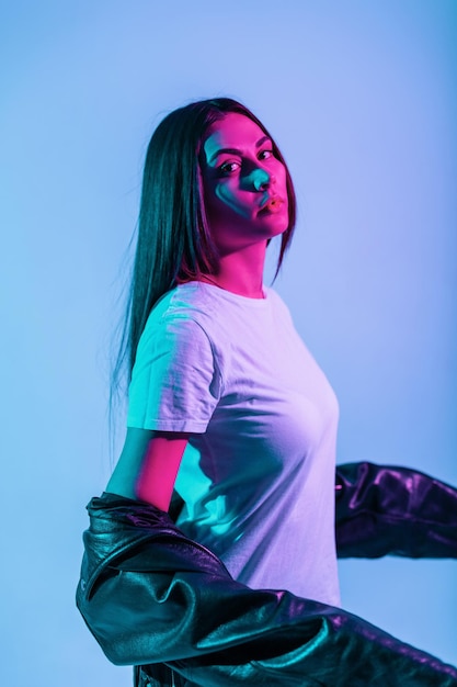 Chica guapa de moda con una cara linda con ropa de moda con una chaqueta negra de cuero y una camiseta blanca posando en el estudio con luces rosas y azules. Retrato creativo de una mujer hermosa