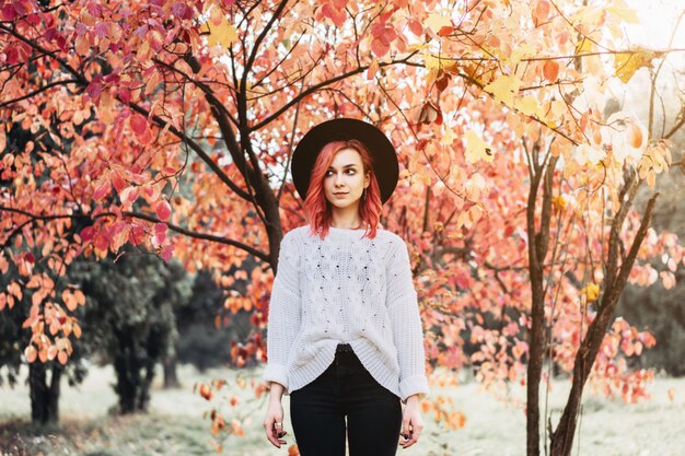 Chica guapa con el pelo rojo y sombrero caminando en el parque, tiempo de otoño.