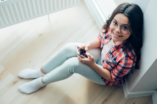 Chica guapa positiva sentada junto a la pared en el suelo con un teléfono inteligente en las manos y mirando hacia arriba con una sonrisa de satisfacción