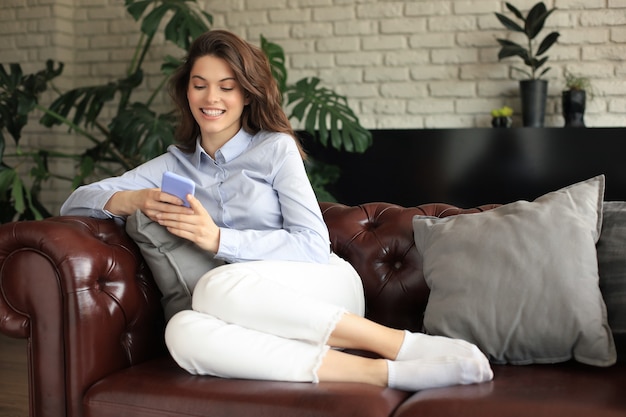 Chica guapa con su teléfono inteligente en el sofá en casa en la sala de estar.